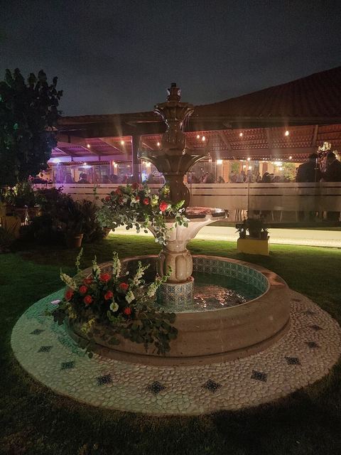 Fuente de cantera decorada con flores, iluminada de noche, con el salón al fondo