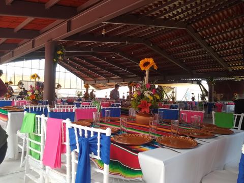 Mesa larga montada con camino de sarape, vajilla de barro y moños de colores en las sillas